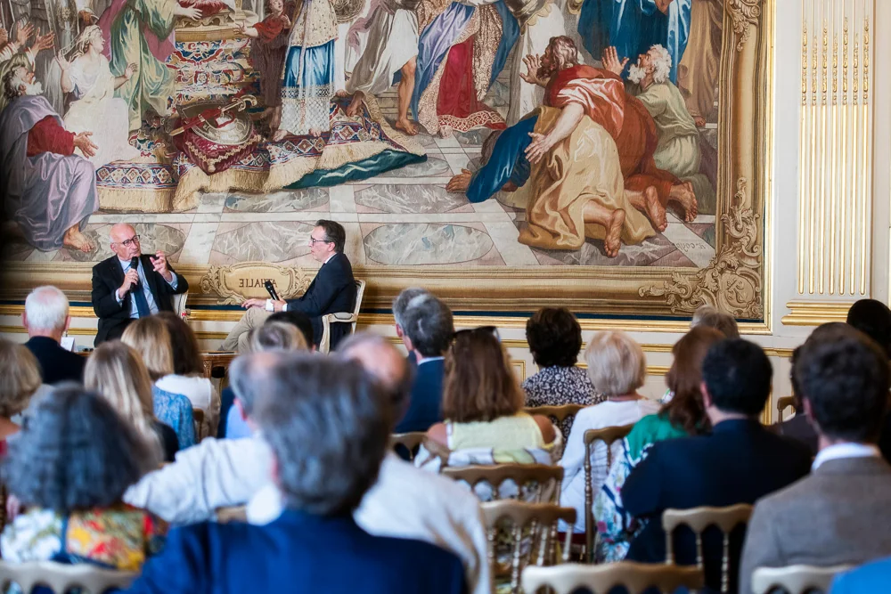 Two speakers talk in front of an audience and a tapestry.
