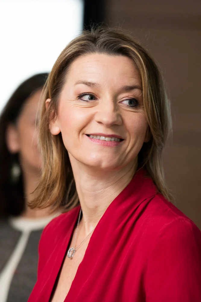 Portrait of an attendee in a red jacket beside a window.