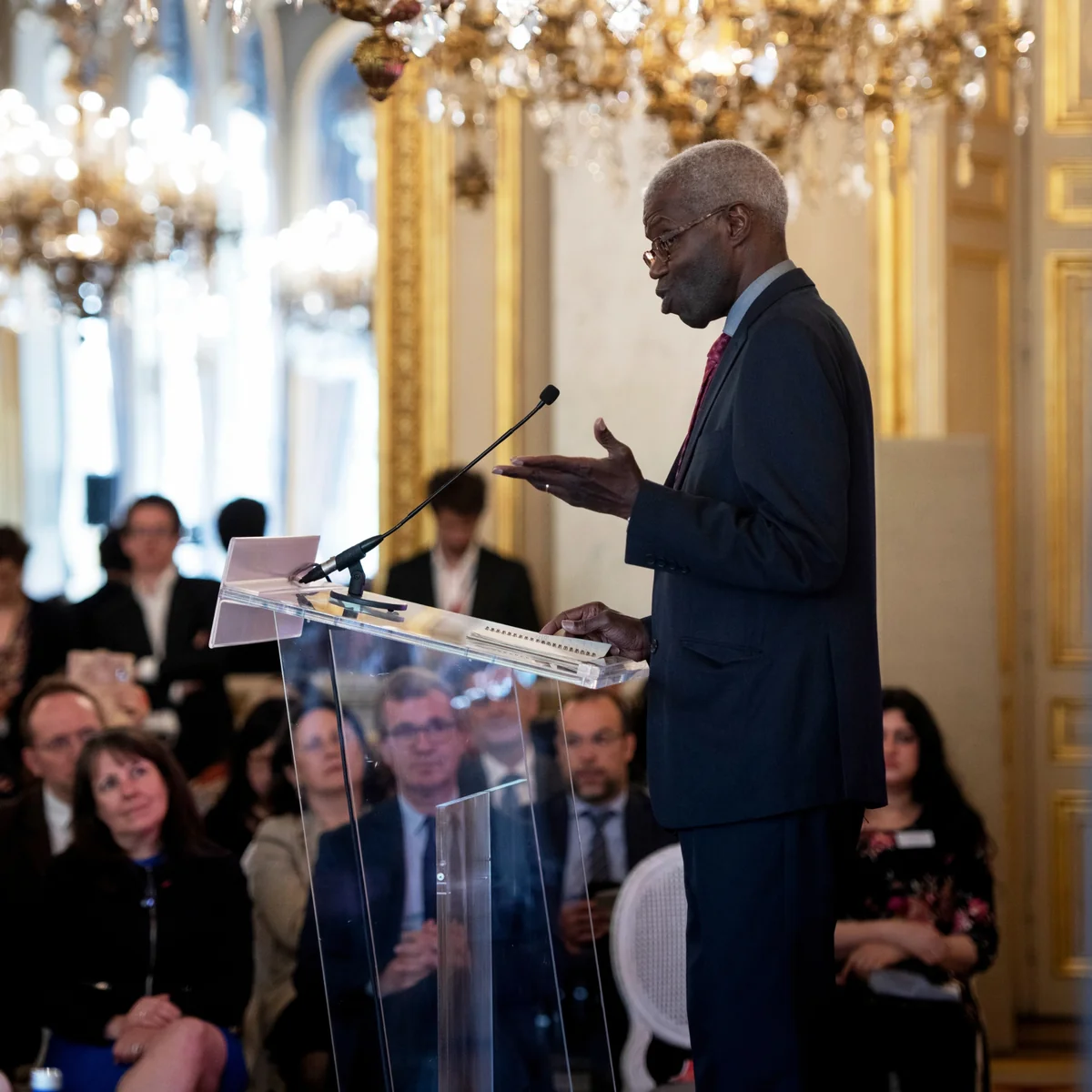 A speaker at a lectern in a room with gilded chandeliers.